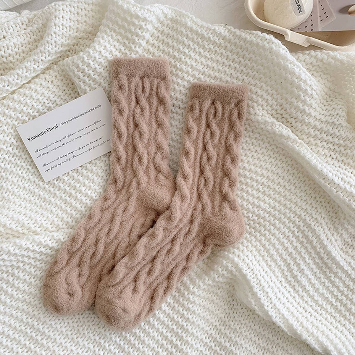 Pair of beige knitted socks on a textured white surface with a card.