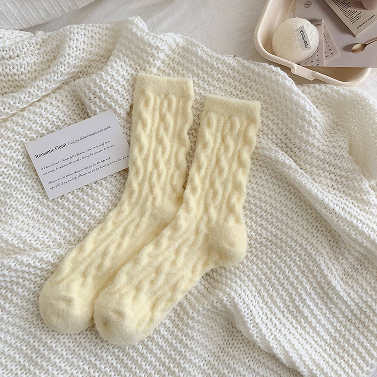 Pair of cream-colored knitted socks on a textured white surface with a card and small container in the background.