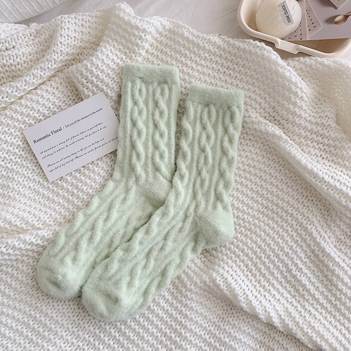Pair of light green cable knit socks on a textured white surface with a card beside them.