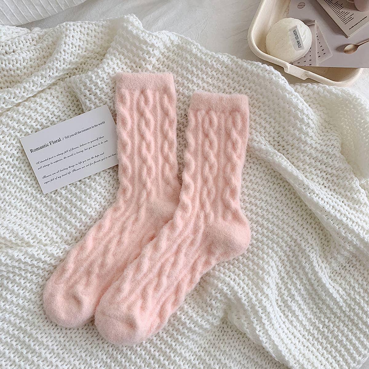 Pair of pink knitted socks on a textured white surface with a card and small container in the background.