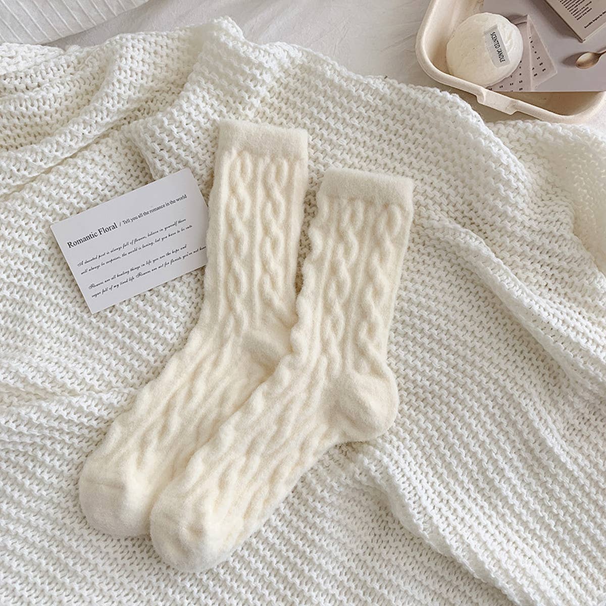Pair of white cable knit socks on a textured surface with a card beside them.