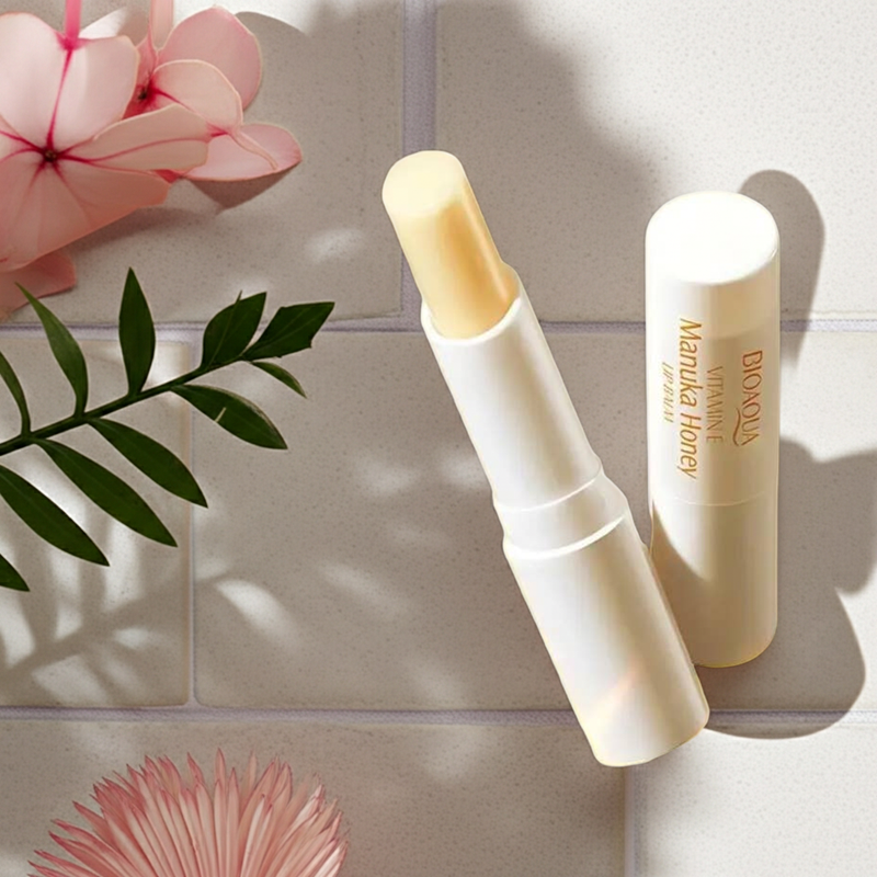 Lip balm tube on a light surface with pink flowers and green leaves in the background