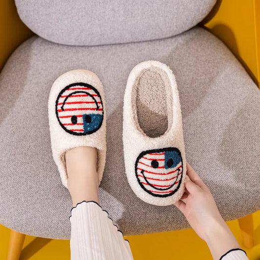 Foot on a grey chair wearing white furry slippers with a smiley face with the style of an American flag