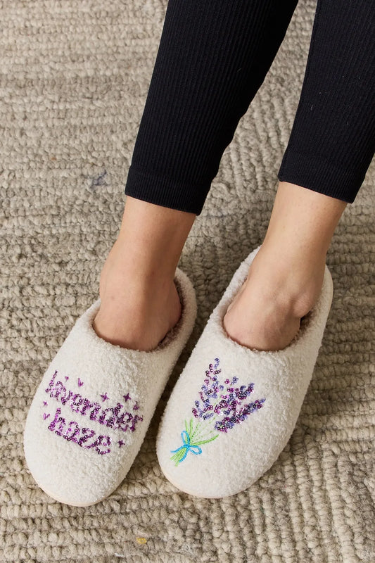 White furry slippers with purple shimmery writing of lavender haze on one slipper and lavender flowers on the other slipper