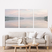 Three-panel misty lake canvas set with blush and pale blue sky, pine-covered shores, and mirror-still water with white couch below panels