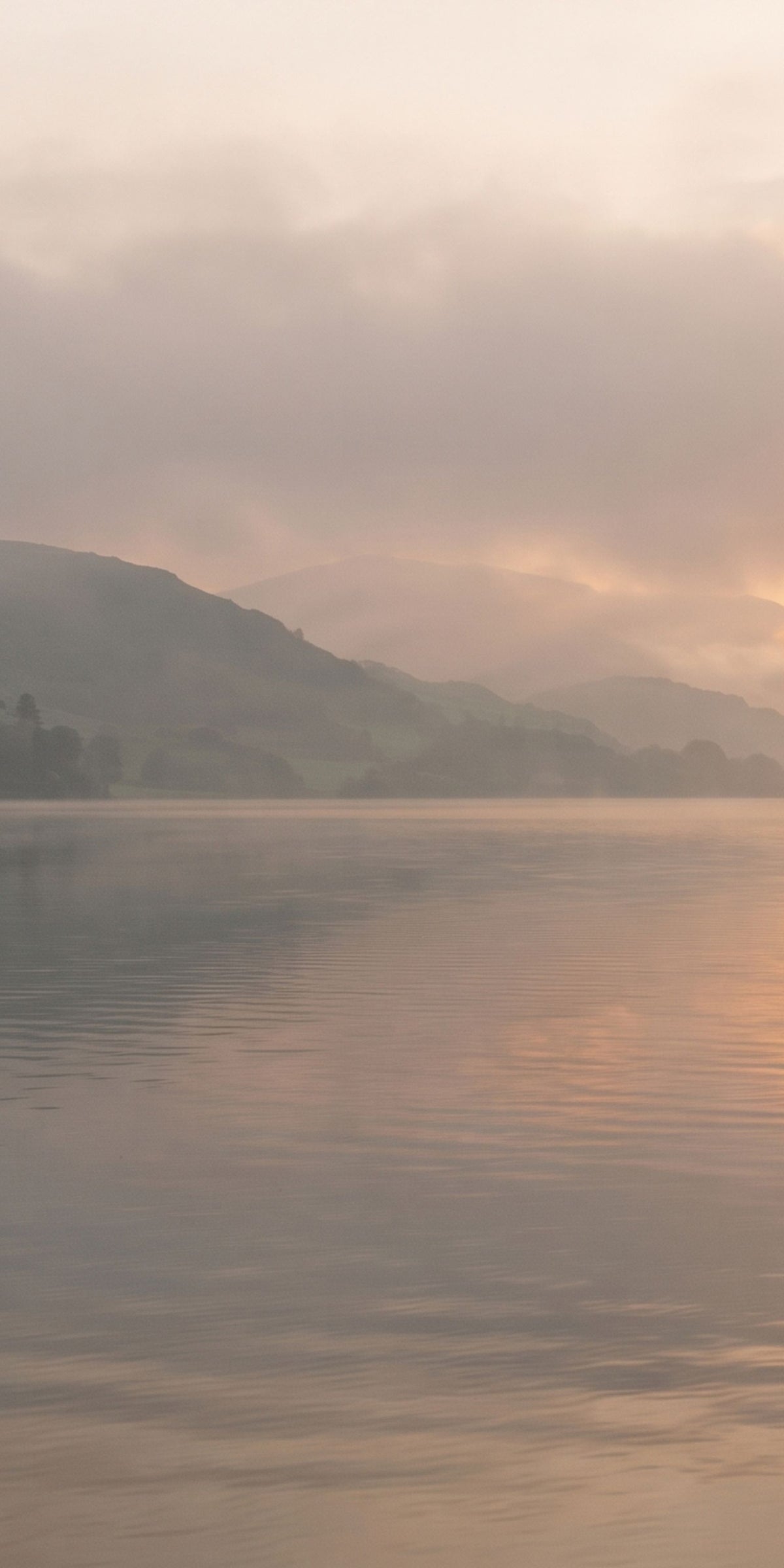 A misty lake landscape at sunrise or sunset, with soft golden light reflecting on calm water and hazy mountains in the background.
