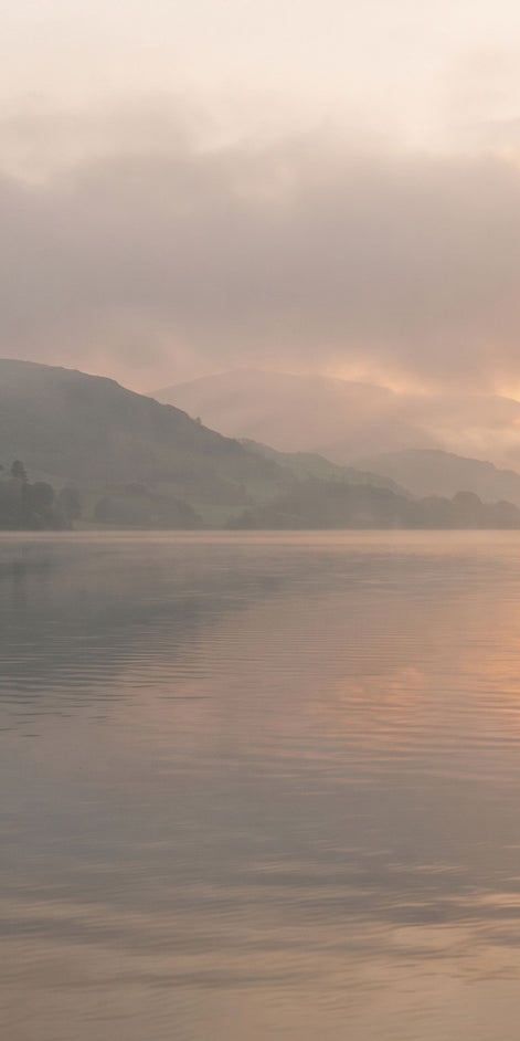 A misty lake landscape at sunrise or sunset, with soft golden light reflecting on calm water and hazy mountains in the background.