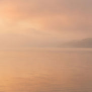 A serene landscape scene of calm water under a soft, misty sky with warm peach and orange light reflecting on the surface.