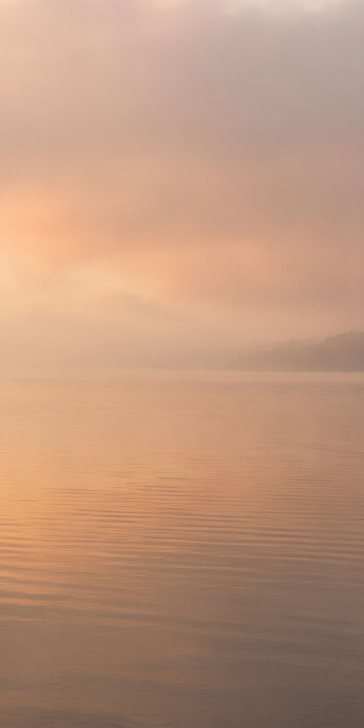 A serene landscape scene of calm water under a soft, misty sky with warm peach and orange light reflecting on the surface.
