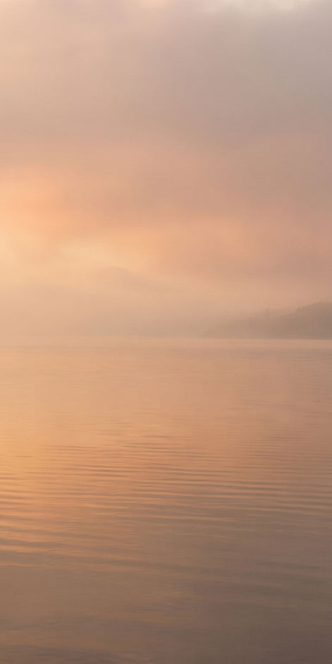 A serene landscape scene of calm water under a soft, misty sky with warm peach and orange light reflecting on the surface.
