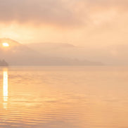 A serene landscape print of a calm lake at sunrise, featuring soft golden-orange clouds, a low sun reflecting on the water, and distant misty mountains.