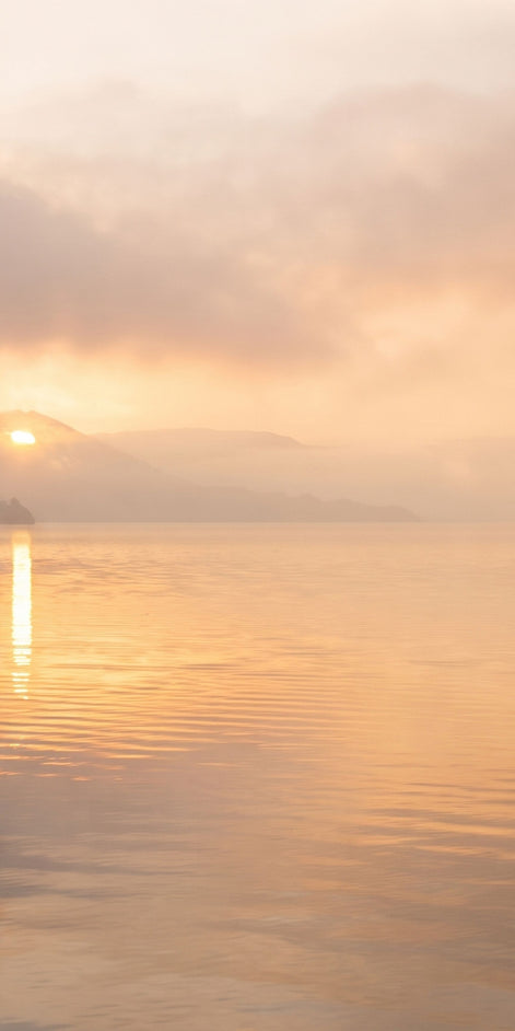 A serene landscape print of a calm lake at sunrise, featuring soft golden-orange clouds, a low sun reflecting on the water, and distant misty mountains.